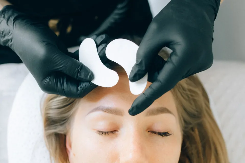 Beauty therapist in black gloves placing protective pads above a customer's eyebrows before a brow treatment