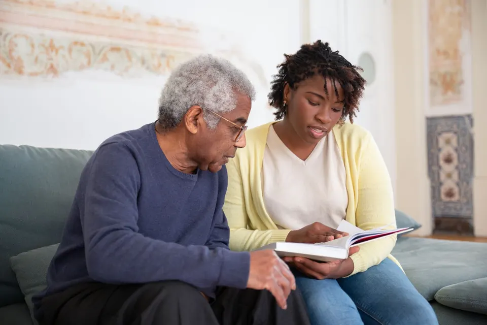 Older man and a carer reading a book together on a sofa at home
