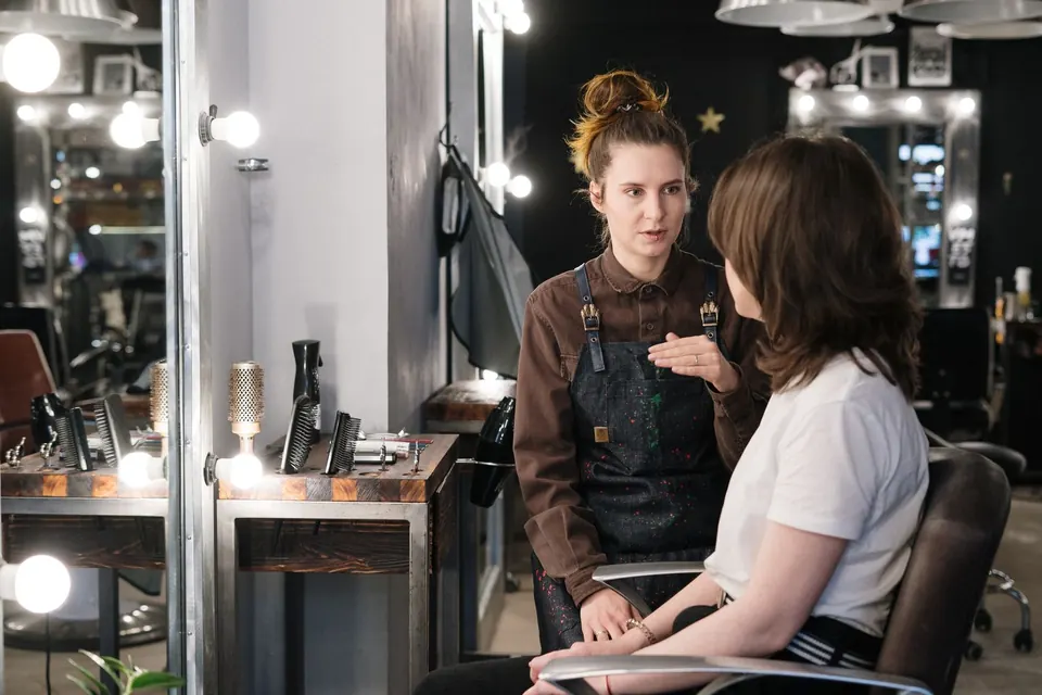 Hair colourist talking to a seated customer at a mirror station before a colour service