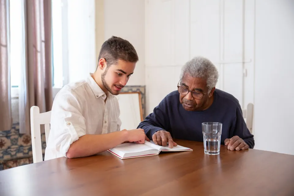 Young carer and an older man reading a book together at a table at home