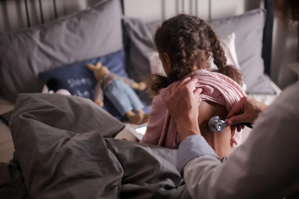 Doctor listening to a child's back with a stethoscope while the child sits on a bed at home
