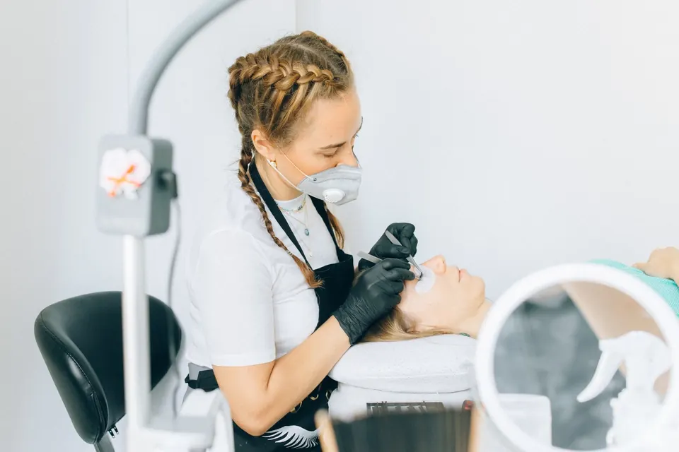 Lash technician applying eyelash extensions to a customer lying on a treatment bed in a bright studio