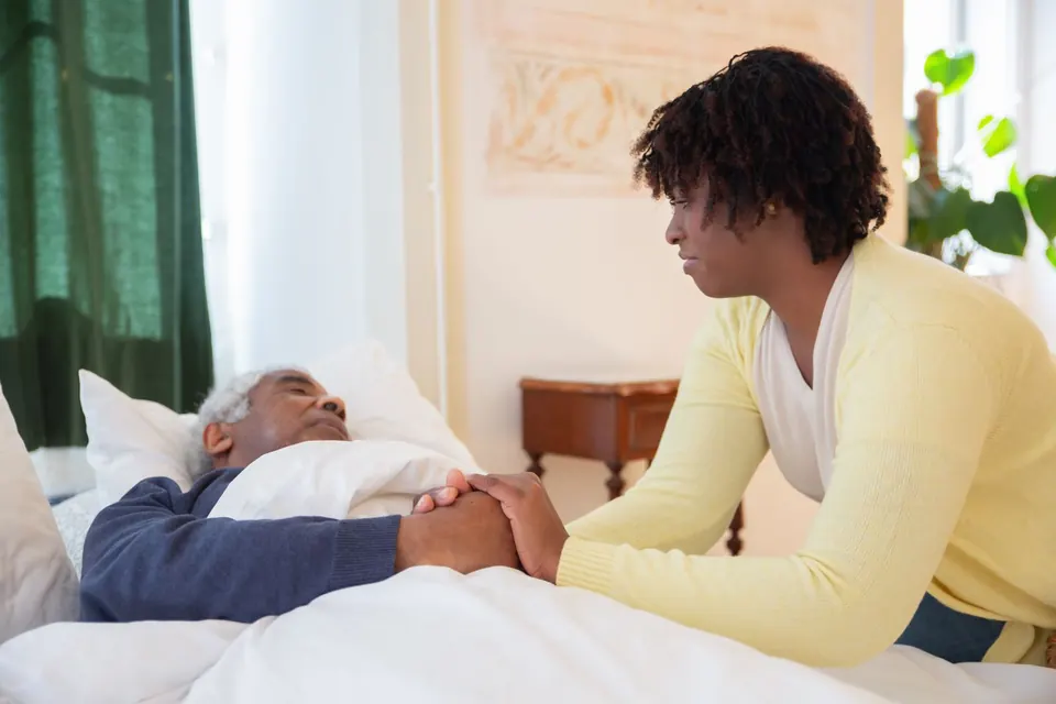 Carer holding the hand of an older man resting in bed at home